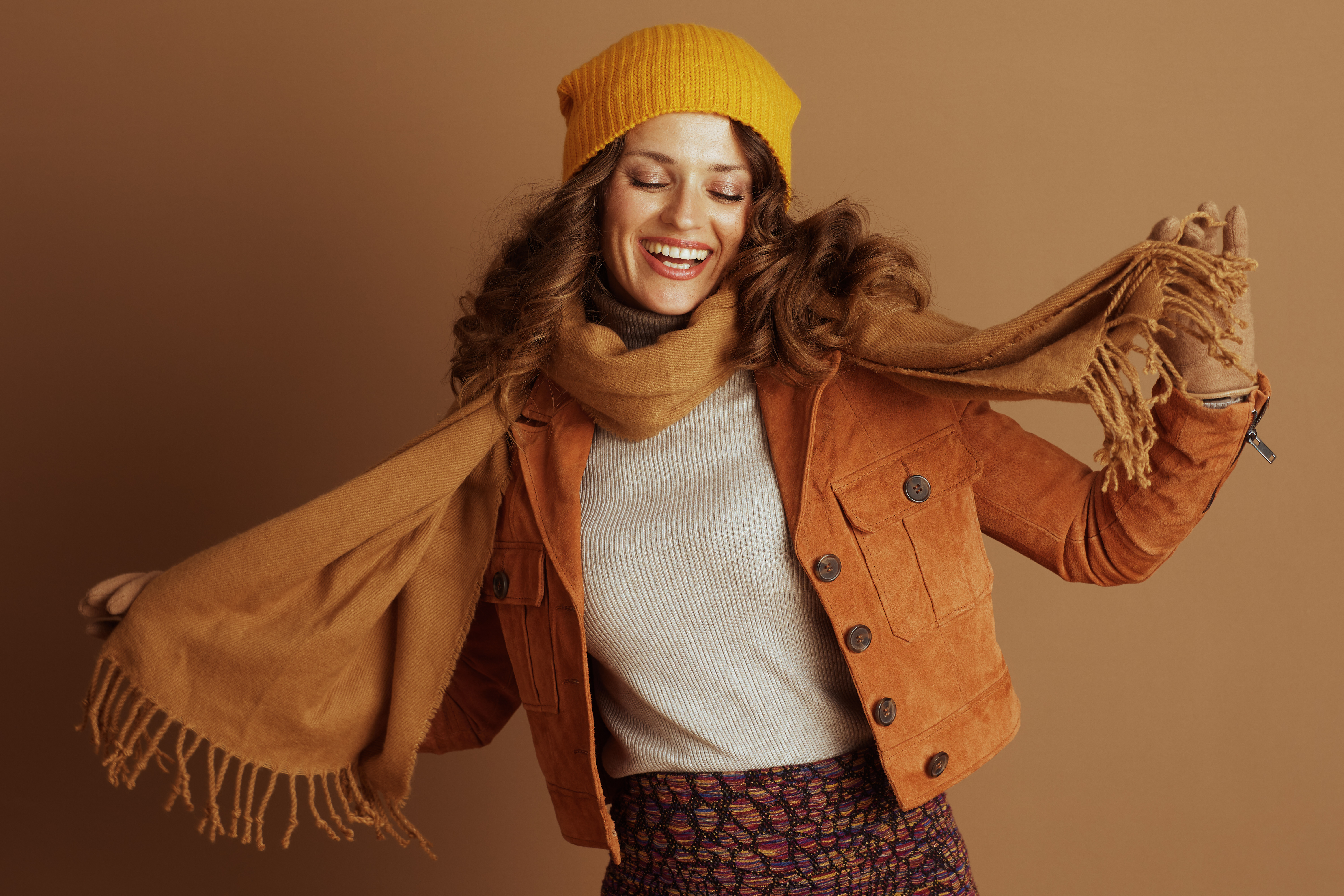 A joyful woman with curly brown hair, wearing a yellow knit beanie, orange suede jacket, and patterned skirt, smiles brightly, eyes closed, playfully gesturing with her tan scarf.